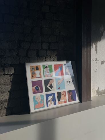 A framed artwork with twelve colorful illustrations rests against an exposed brick wall. Each illustration appears to depict a different subject, including a hand holding a cherry, a figure holding a fan, and a woman with a hat. The lighting creates shadows on the brickwork, adding depth to the scene.