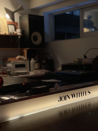 A dimly lit interior space featuring a music studio setup with audio equipment like speakers and a mixing console. The foreground has a prominent illuminated sign with the text 'JOIN WITH US'. The room appears cozy with a warm, inviting atmosphere, likely set up for creative work or a casual gathering.