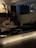 A dimly lit interior space featuring a music studio setup with audio equipment like speakers and a mixing console. The foreground has a prominent illuminated sign with the text 'JOIN WITH US'. The room appears cozy with a warm, inviting atmosphere, likely set up for creative work or a casual gathering.