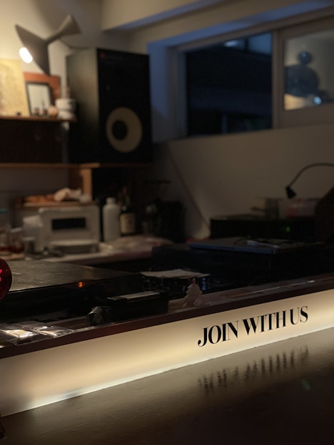 A dimly lit interior space featuring a music studio setup with audio equipment like speakers and a mixing console. The foreground has a prominent illuminated sign with the text 'JOIN WITH US'. The room appears cozy with a warm, inviting atmosphere, likely set up for creative work or a casual gathering.