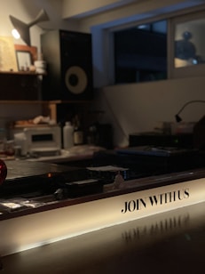 A dimly lit interior space featuring a music studio setup with audio equipment like speakers and a mixing console. The foreground has a prominent illuminated sign with the text 'JOIN WITH US'. The room appears cozy with a warm, inviting atmosphere, likely set up for creative work or a casual gathering.