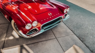 A classic red Corvette with a shiny surface is parked on a pavement. The car's chrome details and whitewall tires are also visible. The pavement is made of concrete tiles and there is a road with a rougher texture next to it.