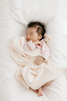 A sleeping baby is swaddled in a soft, pale pink blanket. The baby is wearing a pink and white striped outfit and has dark hair. The background is a white, fluffy surface, adding to the cozy feel.