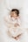 A sleeping baby is swaddled in a soft, pale pink blanket. The baby is wearing a pink and white striped outfit and has dark hair. The background is a white, fluffy surface, adding to the cozy feel.