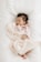 A sleeping baby is swaddled in a soft, pale pink blanket. The baby is wearing a pink and white striped outfit and has dark hair. The background is a white, fluffy surface, adding to the cozy feel.