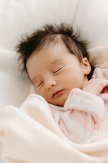 A peaceful sleeping baby wrapped in a soft, light-colored blanket. The baby has dark hair and wears a pink outfit.