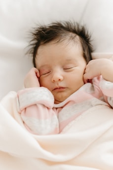 A sleeping baby with soft, dark hair is wrapped snugly in a creamy white blanket. The baby is wearing a pink and white striped outfit and appears peaceful and serene.
