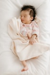 A newborn baby peacefully sleeps wrapped in a soft, pale pink blanket. The baby is wearing a pink and gray striped onesie, and is positioned on a white, cozy surface. The baby has dark hair and is lying with a relaxed expression.