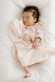A cozy baby girl wearing a soft pastel pink onesie, sitting on a fluffy white blanket.