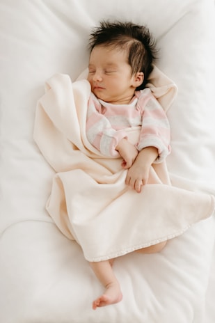 A newborn baby peacefully sleeps wrapped in a soft, pale pink blanket. The baby is wearing a pink and gray striped onesie, and is positioned on a white, cozy surface. The baby has dark hair and is lying with a relaxed expression.