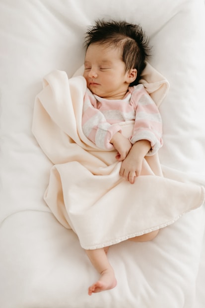 A newborn baby peacefully sleeps wrapped in a soft, pale pink blanket. The baby is wearing a pink and gray striped onesie, and is positioned on a white, cozy surface. The baby has dark hair and is lying with a relaxed expression.