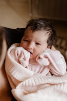 A newborn baby is wrapped snugly in a soft, knitted pink blanket. The baby is wearing a pink onesie and has chubby cheeks and dark hair, with hands raised close to its mouth.