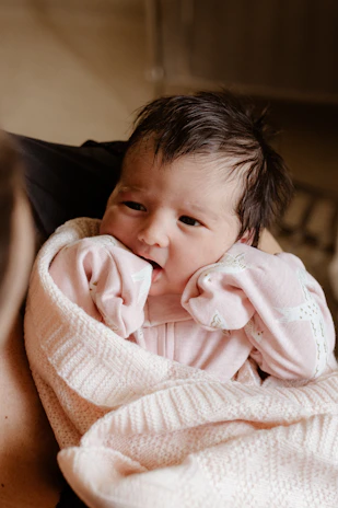 A smiling newborn wrapped in a soft pink swaddle, lying on a fluffy white blanket.