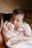 A newborn baby is wrapped snugly in a soft, knitted pink blanket. The baby is wearing a pink onesie and has chubby cheeks and dark hair, with hands raised close to its mouth.