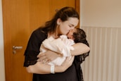 A woman lovingly holds and kisses a newborn baby wrapped in a soft blanket. They are indoors with a wooden door and light-colored walls creating a warm and intimate setting.