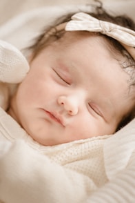 A peaceful, sleeping baby with a headband rests comfortably. The baby wears soft, warm clothing in neutral shades and has delicate features, creating a serene and tender atmosphere.