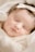 A peaceful, sleeping baby with a headband rests comfortably. The baby wears soft, warm clothing in neutral shades and has delicate features, creating a serene and tender atmosphere.