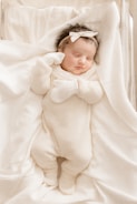 A newborn baby peacefully sleeps wrapped in a soft white blanket, wearing a white knitted onesie and mittens. The baby has a white bow headband and lies on a gently textured surface.