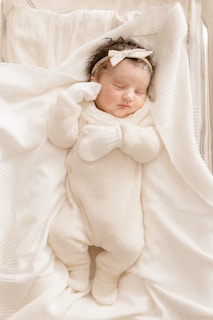 A newborn baby peacefully sleeps wrapped in a soft white blanket, wearing a white knitted onesie and mittens. The baby has a white bow headband and lies on a gently textured surface.