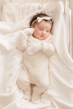 A newborn baby peacefully sleeps wrapped in a soft white blanket, wearing a white knitted onesie and mittens. The baby has a white bow headband and lies on a gently textured surface.