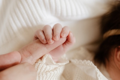 A small infant's hand gently grasping an adult's finger, surrounded by a soft textured fabric. The scene conveys a sense of tenderness and care.