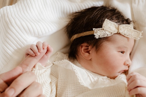 A newborn baby with dark hair is lying on a soft, textured white blanket, wearing a knitted outfit and a delicate lace headband with a bow. The baby is gently holding onto an adult's finger with its small hand.