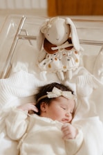 A newborn baby is lying down, wrapped in a white blanket, and wears a cream-colored outfit with a lace headband. Beside the baby, there's a soft toy bunny dressed in a cute polka-dotted dress placed inside a clear plastic crib.