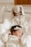 A newborn baby is lying down, wrapped in a white blanket, and wears a cream-colored outfit with a lace headband. Beside the baby, there's a soft toy bunny dressed in a cute polka-dotted dress placed inside a clear plastic crib.