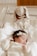 A newborn baby is lying down, wrapped in a white blanket, and wears a cream-colored outfit with a lace headband. Beside the baby, there's a soft toy bunny dressed in a cute polka-dotted dress placed inside a clear plastic crib.