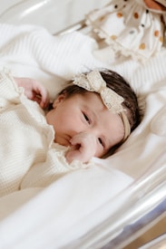 A newborn baby lies peacefully in a hospital bassinet, wrapped in a cream-colored knit blanket. The baby is wearing a delicate lace headband with a bow. The overall setting is calm and soft, with cream and light colors, suggesting a warm and gentle atmosphere.