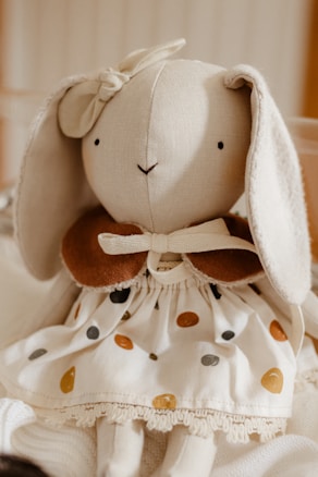 A soft fabric rabbit doll features long floppy ears and a simple, embroidered face. The doll is adorned with a bow on one ear and wears a white dress decorated with multicolored polka dots. A brown collar adds contrast to the outfit.