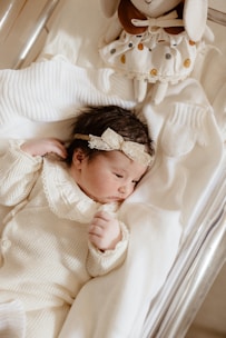 A newborn baby dressed in a cozy, cream-colored knitted outfit lies peacefully on a soft, white blanket. The baby's head is adorned with a delicate lace headband. A plush toy with a polka dot dress is positioned near the baby's head, adding a touch of warmth and charm to the scene.