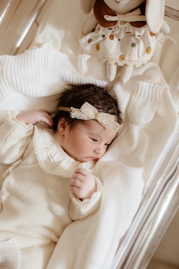 A newborn baby dressed in a cozy, cream-colored knitted outfit lies peacefully on a soft, white blanket. The baby's head is adorned with a delicate lace headband. A plush toy with a polka dot dress is positioned near the baby's head, adding a touch of warmth and charm to the scene.