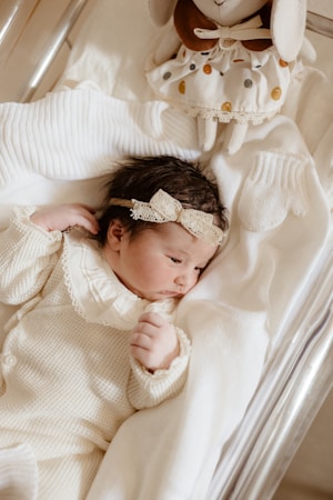 A newborn baby dressed in a cozy, cream-colored knitted outfit lies peacefully on a soft, white blanket. The baby's head is adorned with a delicate lace headband. A plush toy with a polka dot dress is positioned near the baby's head, adding a touch of warmth and charm to the scene.