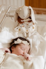 A sleeping baby dressed in neutral-colored clothing lies in a crib. Above the baby, a plush toy resembling a rabbit with long ears and wearing a dress is placed. The atmosphere is soft and serene.