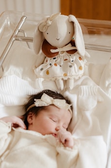 A sleeping baby dressed in neutral-colored clothing lies in a crib. Above the baby, a plush toy resembling a rabbit with long ears and wearing a dress is placed. The atmosphere is soft and serene.