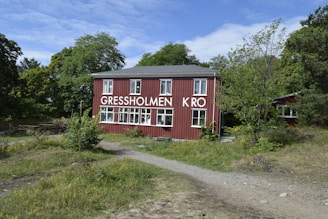 A red wooden building with multiple white-framed windows is surrounded by greenery and trees. The building has the words 'GRESSHOLMEN KRO' displayed prominently on its facade. A dirt pathway leads up to the structure, which is set in a natural setting with grass and small plants in the foreground. The sky is partly cloudy, suggesting a mild day.