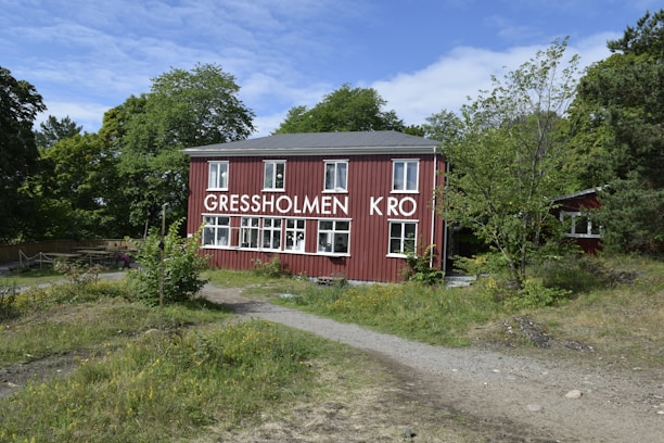 A red wooden building with multiple white-framed windows is surrounded by greenery and trees. The building has the words 'GRESSHOLMEN KRO' displayed prominently on its facade. A dirt pathway leads up to the structure, which is set in a natural setting with grass and small plants in the foreground. The sky is partly cloudy, suggesting a mild day.