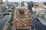 Aerial view of modern urban architecture featuring a prominent building with multiple terraces adorned with greenery. Surrounding the building are other contemporary structures, a river, and various urban features like roads and bridges. The skyline shows a mix of glass and concrete buildings in a bustling city environment.