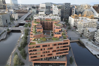 Aerial view of modern urban architecture featuring a prominent building with multiple terraces adorned with greenery. Surrounding the building are other contemporary structures, a river, and various urban features like roads and bridges. The skyline shows a mix of glass and concrete buildings in a bustling city environment.
