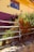A rustic wooden fence borders a garden full of colorful flowers, with a yellow and purple-painted building in the background. The window of the building reflects the clear blue sky, and the wall has two small potted plants affixed near the top. Stones and some vegetation can be seen at the base of the fence.
