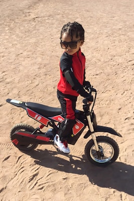 A child wearing a red and black outfit with sunglasses is sitting on a small dirt bike in a sandy area. The child appears confident, with hands on the handlebars and looking towards the side.