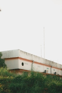 A rectangular building with a flat roof, displaying signs of weathering, stands behind a lush grassy area. The top of the building has railings and antennas protruding. The walls are painted white with an orange trim, and there are small round windows and air conditioning units visible.