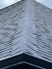 A close-up view of a roof with dark, overlapping shingles creating a textured surface. The shingles appear to be evenly laid with a symmetrical design.