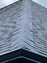 A close-up view of a roof with dark, overlapping shingles creating a textured surface. The shingles appear to be evenly laid with a symmetrical design.