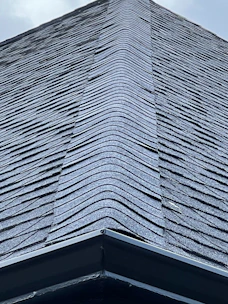 A close-up view of a roof with dark, overlapping shingles creating a textured surface. The shingles appear to be evenly laid with a symmetrical design.