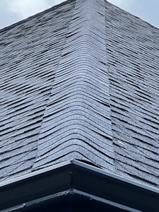 A close-up view of a roof with dark, overlapping shingles creating a textured surface. The shingles appear to be evenly laid with a symmetrical design.