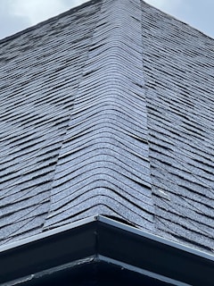 A close-up view of a roof with dark, overlapping shingles creating a textured surface. The shingles appear to be evenly laid with a symmetrical design.
