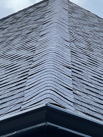 A close-up view of a roof with dark, overlapping shingles creating a textured surface. The shingles appear to be evenly laid with a symmetrical design.
