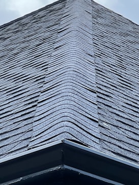 A close-up view of a roof with dark, overlapping shingles creating a textured surface. The shingles appear to be evenly laid with a symmetrical design.
